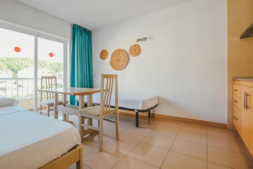 Ein Zimmer mit einem Bett, einem Tisch, einem Stuhl und einem kleinen Schreibtisch. An den Wänden hängen drei gewebte Wanddekorationen und ein Fenster mit Vorhängen.