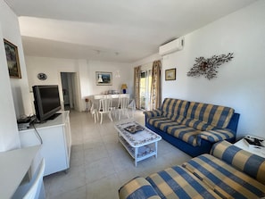 Sala de estar com sofá, televisão, área de refeições com cadeiras e mesa, e um objeto decorativo na parede.