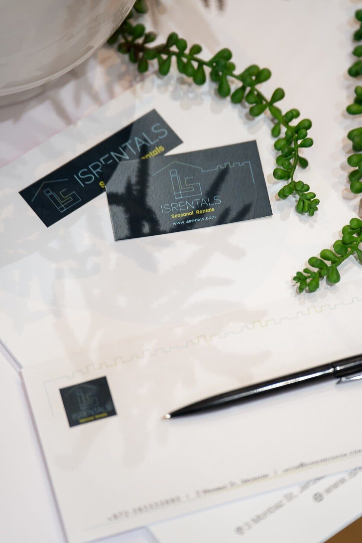 Des cartes de visite et un stylo sont posés sur une table à la surface blanche, entourée de feuillage vert.
