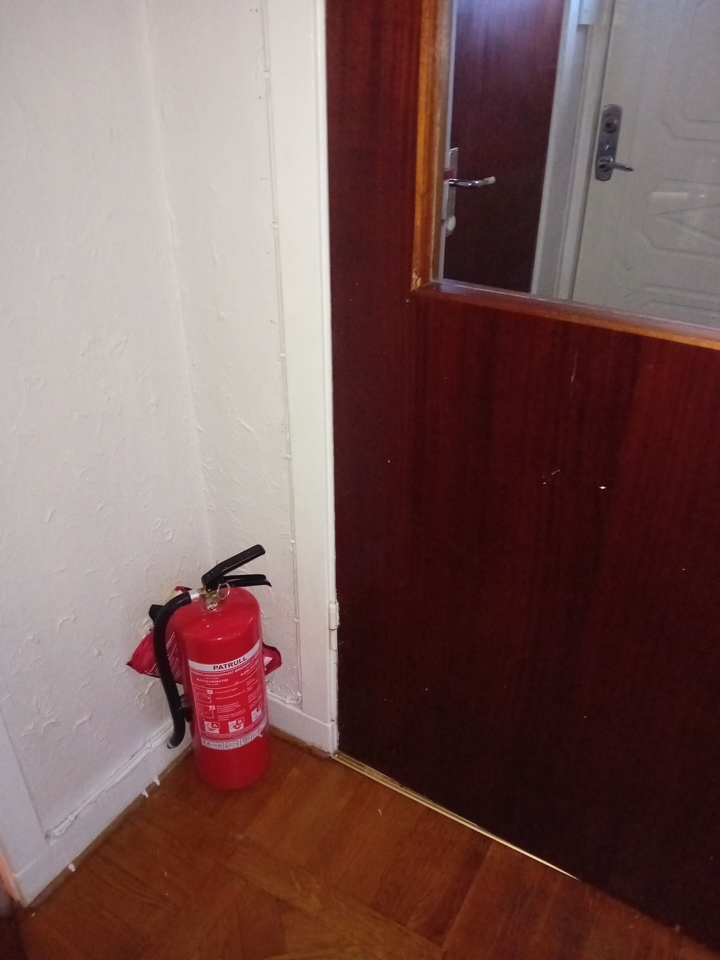 Ein roter Feuerlöscher ist an einer weißen Wand neben einer Holztür angebracht.