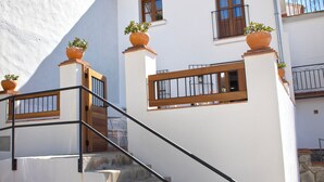 A white building with a staircase leading to a wooden door. There are potted plants on the steps and railings.