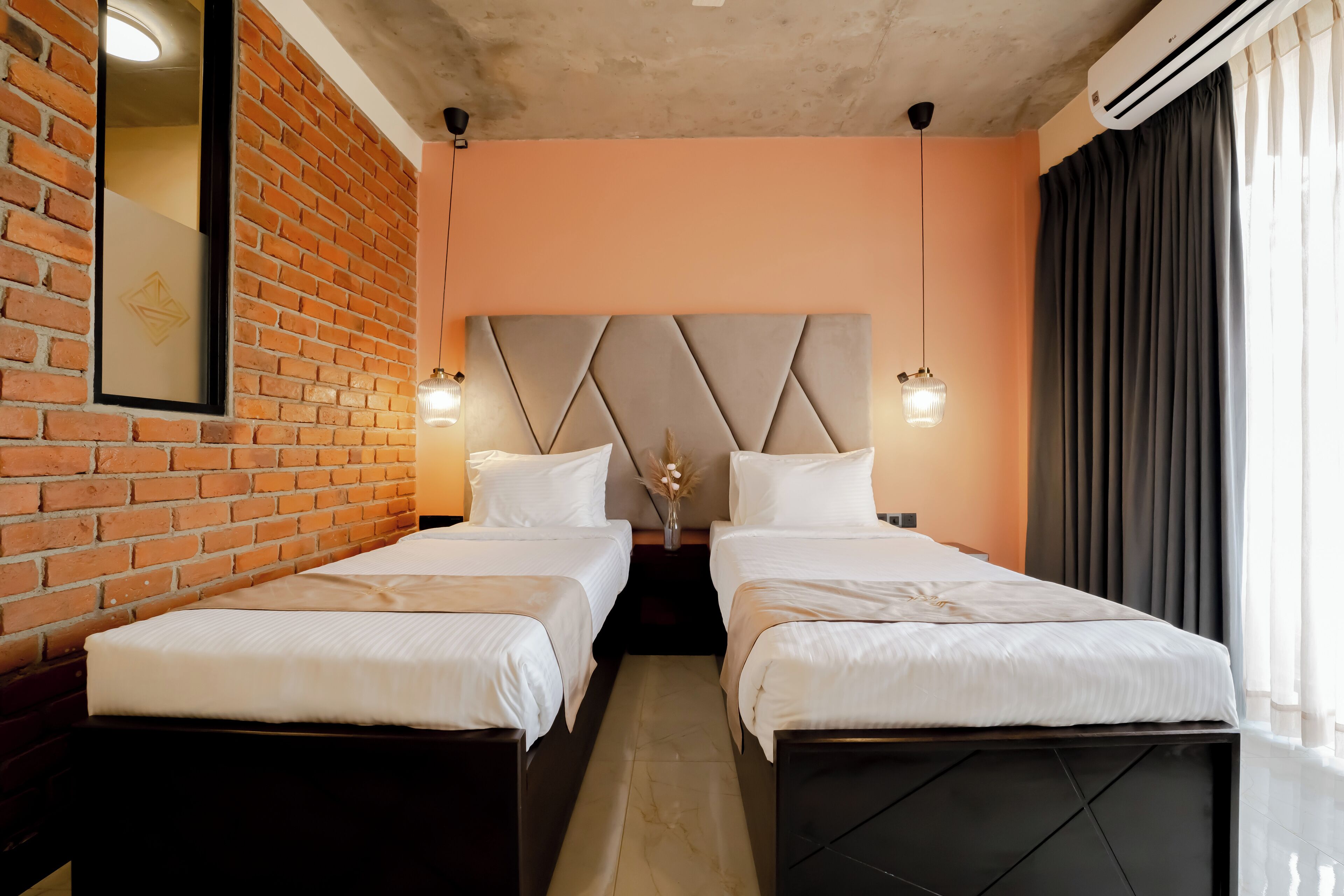 Une chambre d’hôtel avec deux lits, un mur en briques, une tête de lit moderne et une grande fenêtre avec des rideaux.