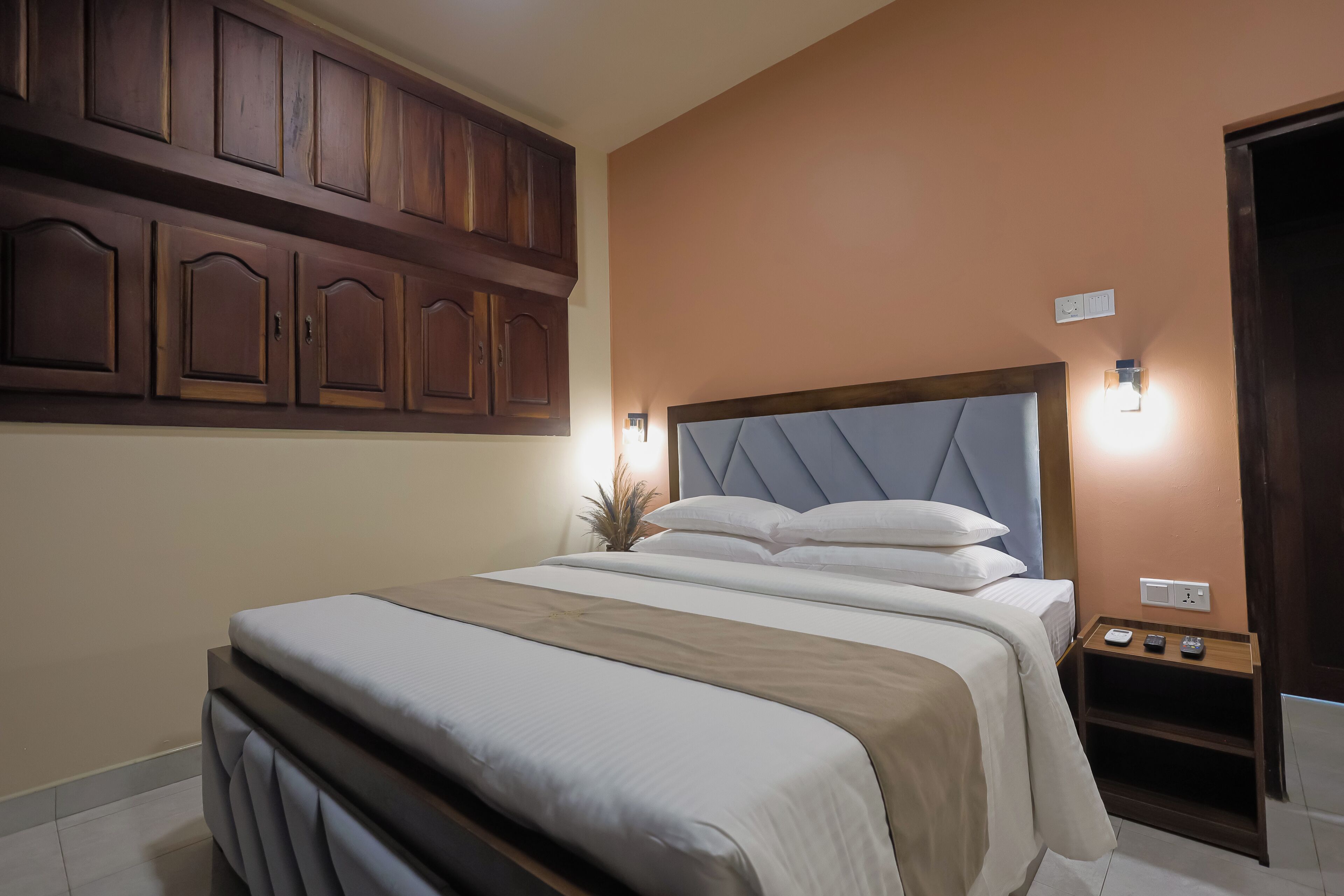A neatly made bed with a headboard, a bedside table with a lamp, and wooden cabinets above the bed.