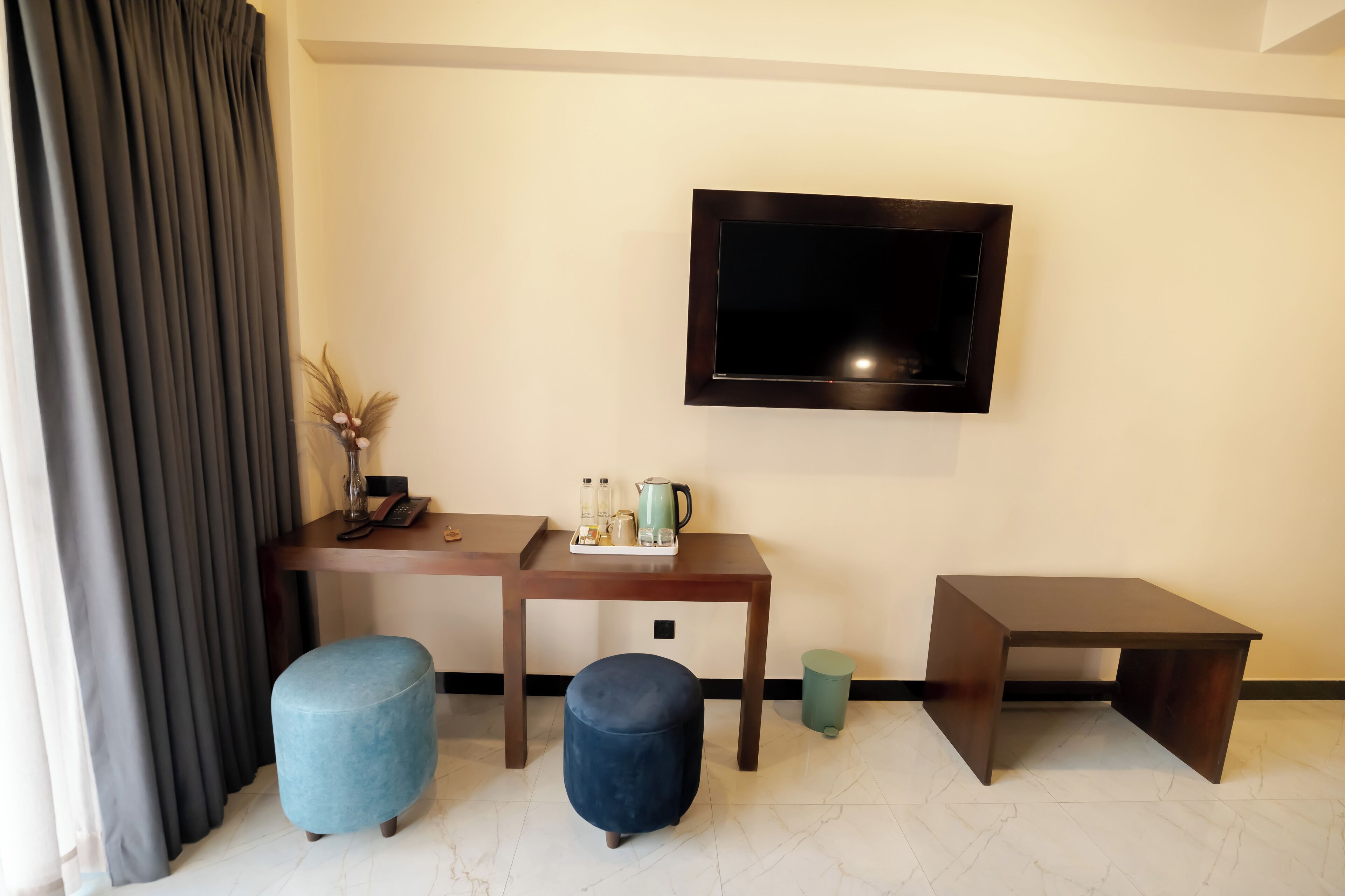 A hotel room with a flat-screen TV mounted on the wall, a wooden desk with a tray of items, two ottomans, and a small table.