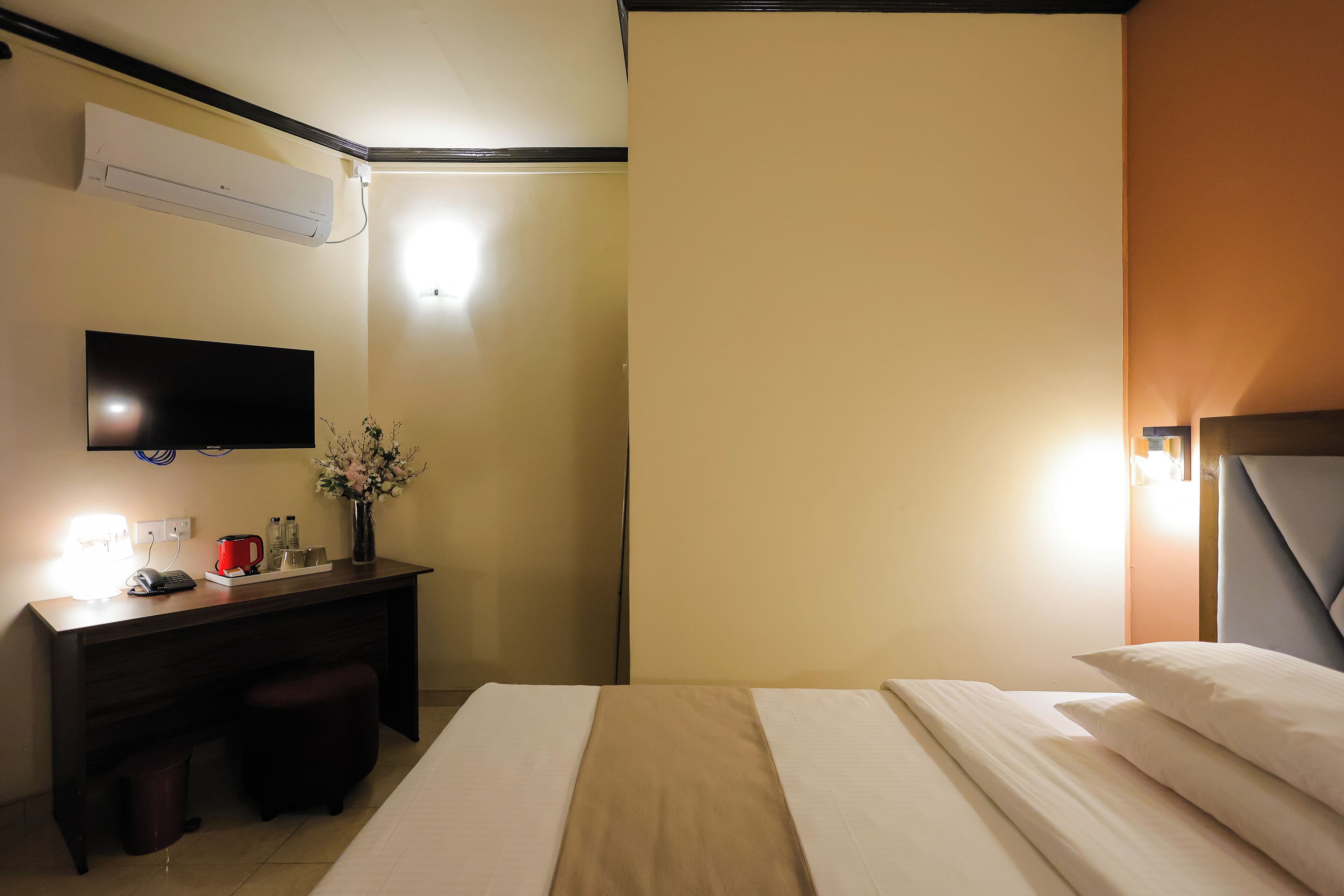 A hotel room with a bed, a desk with a lamp, a TV, and a wall-mounted air conditioner.