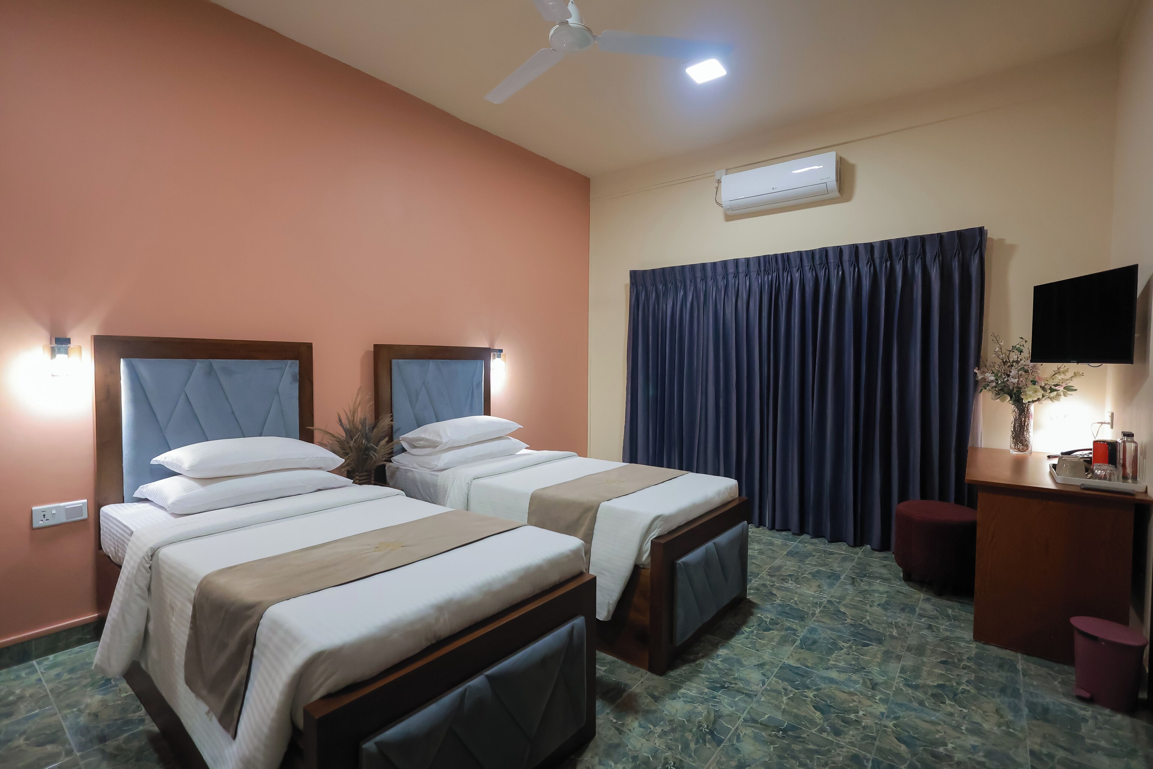 Ein Hotelzimmer mit zwei Betten, einem Schreibtisch, einem Flachbildfernseher, einem Deckenventilator und einer Klimaanlage.