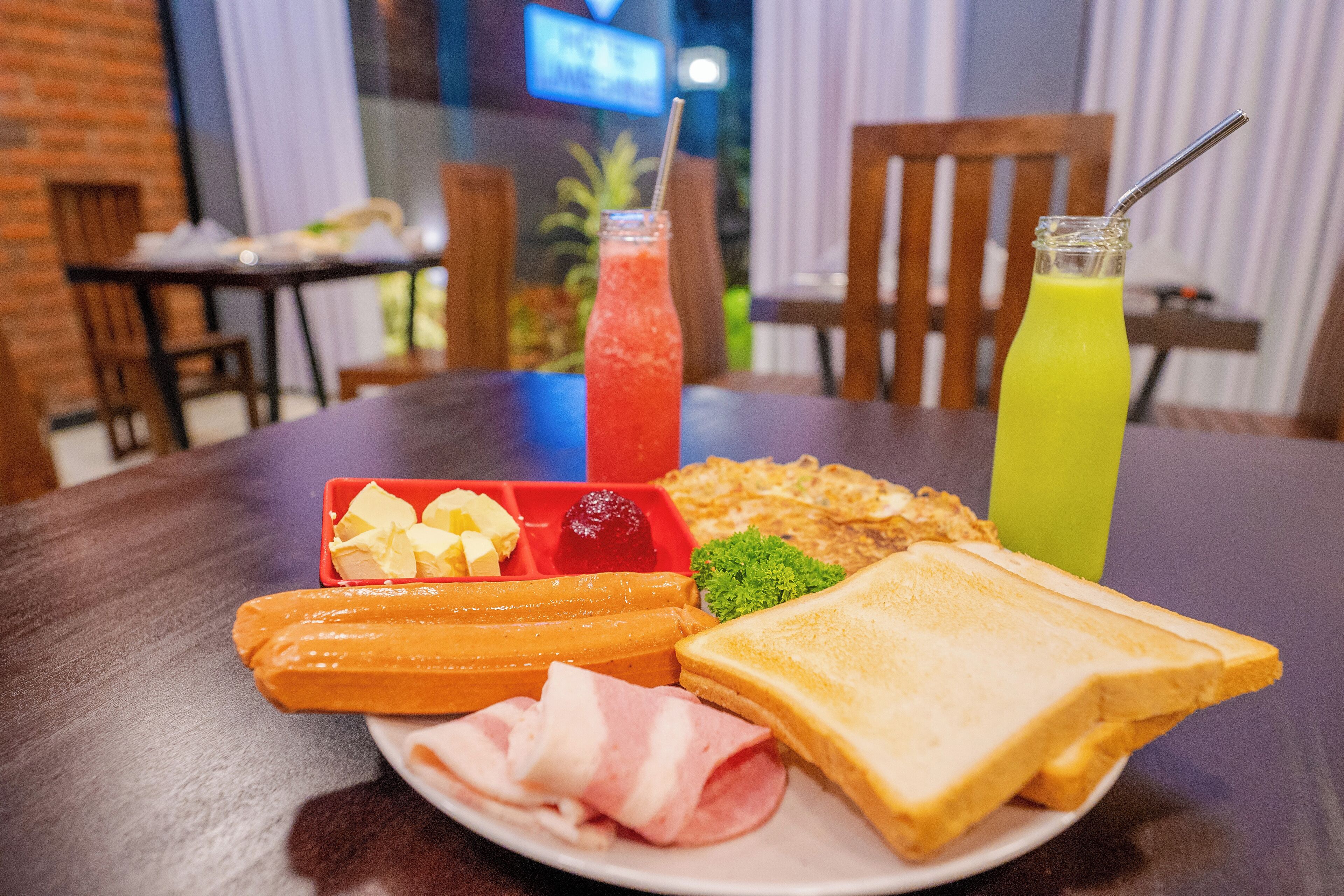 Un plato de desayuno con tostadas, jamón, queso y una ensalada de frutas, junto con dos bebidas sobre una mesa en un comedor.