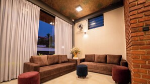Una sala moderna con dos sofás marrones, un pouf rojo y otro pouf oscuro. Hay una ventana con cortinas blancas, un ventilador de pared y una pared de ladrillo decorativa.