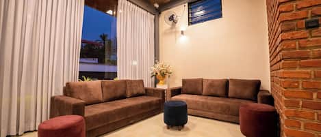 Ein modernes Wohnzimmer mit zwei braunen Sofas, einem roten Hocker und einem dunklen Ottomanen. Ein Fenster mit weißen Vorhängen, ein Wandventilator und eine Ziegelwand sind zu sehen.