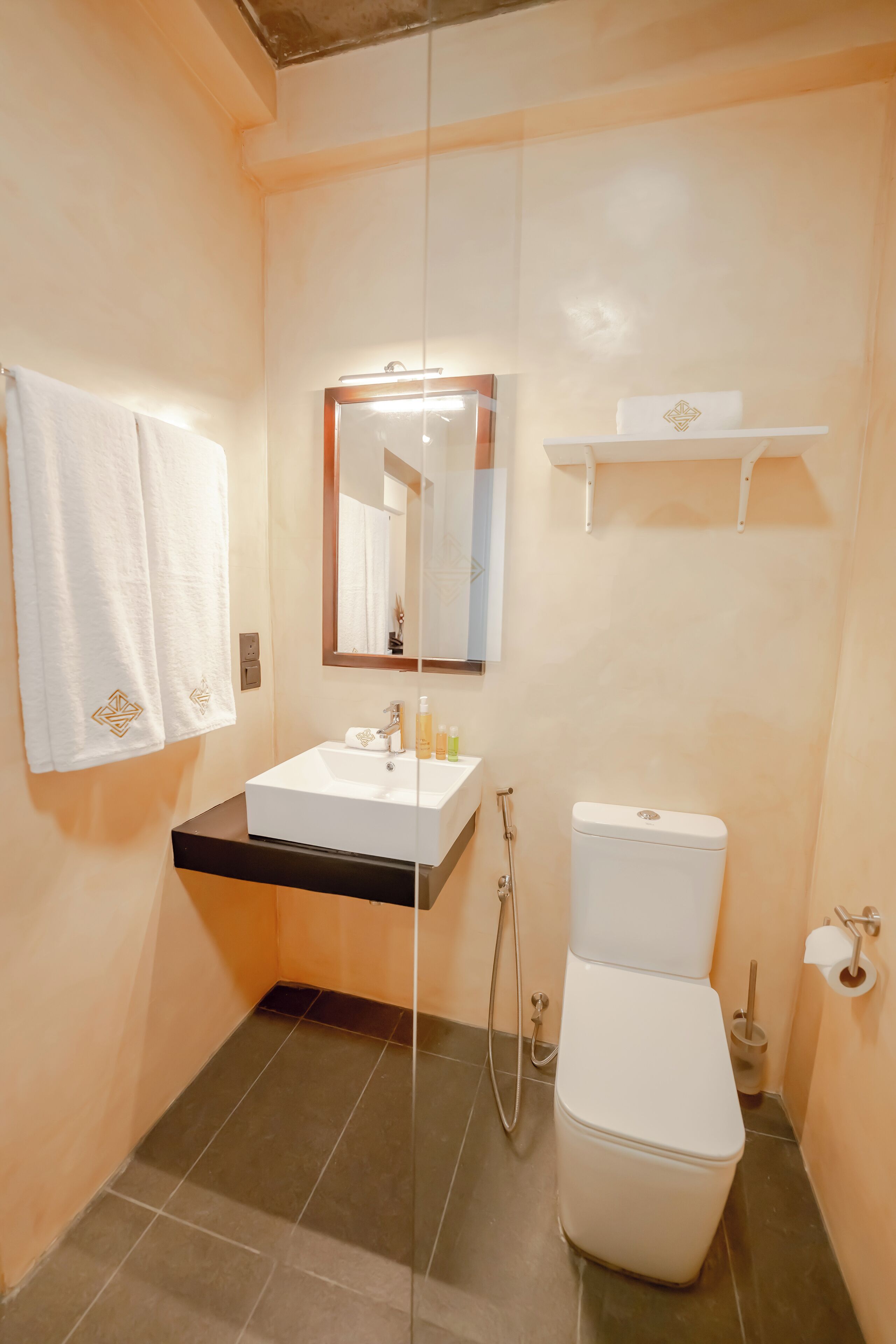 A bathroom with a white sink, mirror, and toilet. There are towels hanging on the wall and a shelf above the toilet.