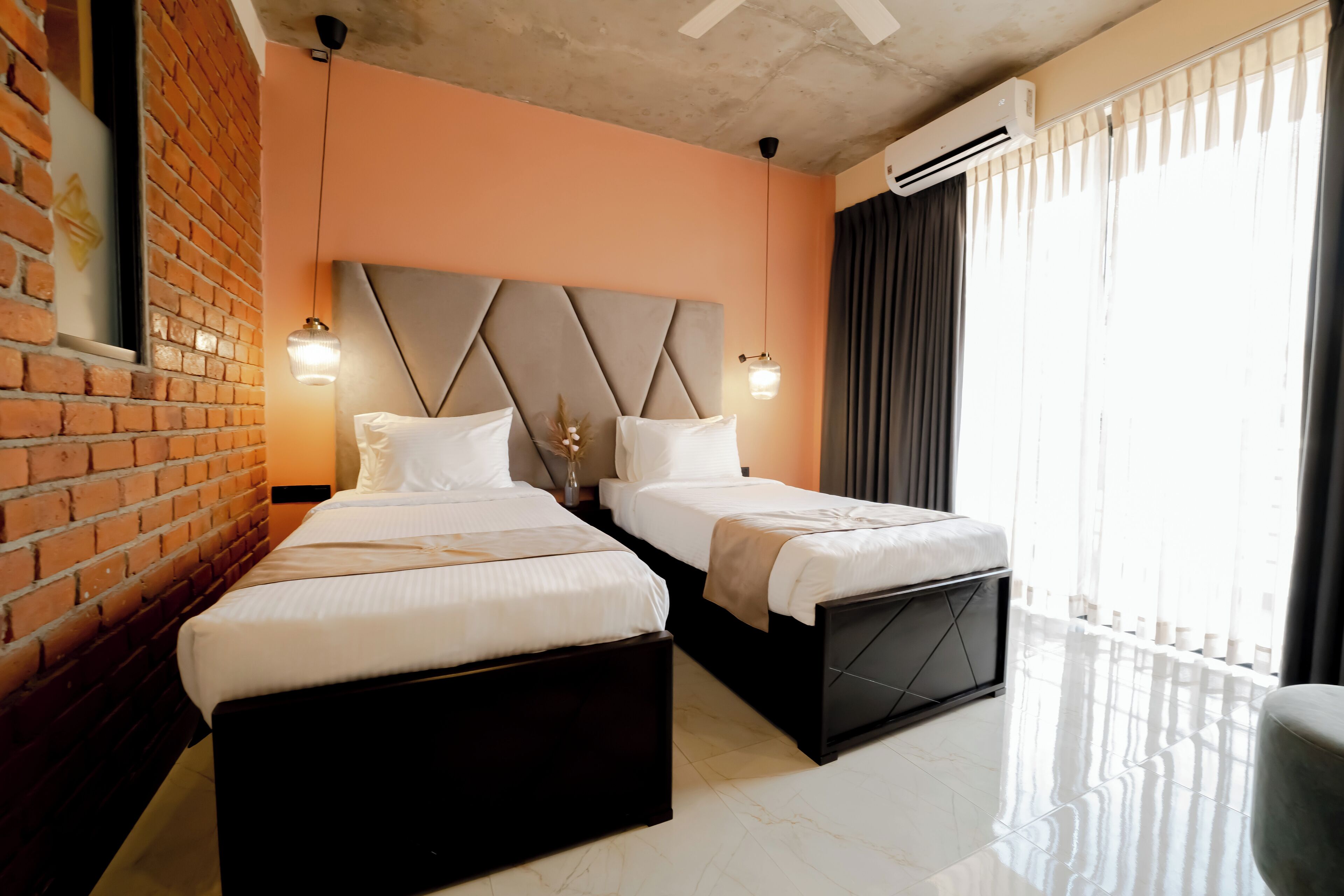 A hotel room with two beds, a brick wall, a modern headboard, and a large window with curtains.