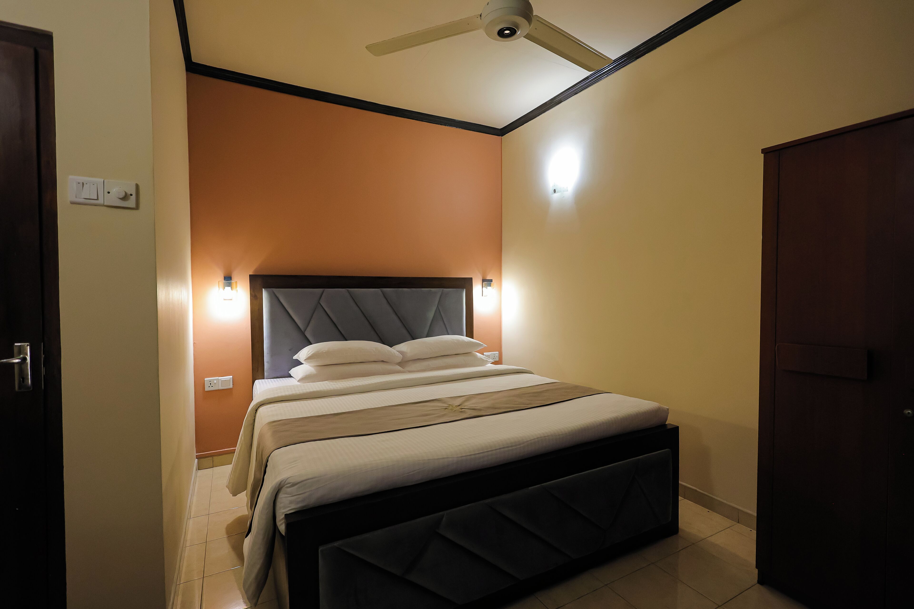 Een keurig opgemaakt bed met hoofdbord, een plafondventilator en een wandlamp.