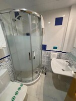 Un baño con ducha envuelta en vidrio, un lavabo blanco y un inodoro con símbolos verdes.