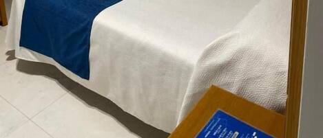 Ein Einzelbett mit weißblauer Bettdecke, ein an der Wand befestigtes Fernsehgerät und ein Nachttisch mit Lampe.