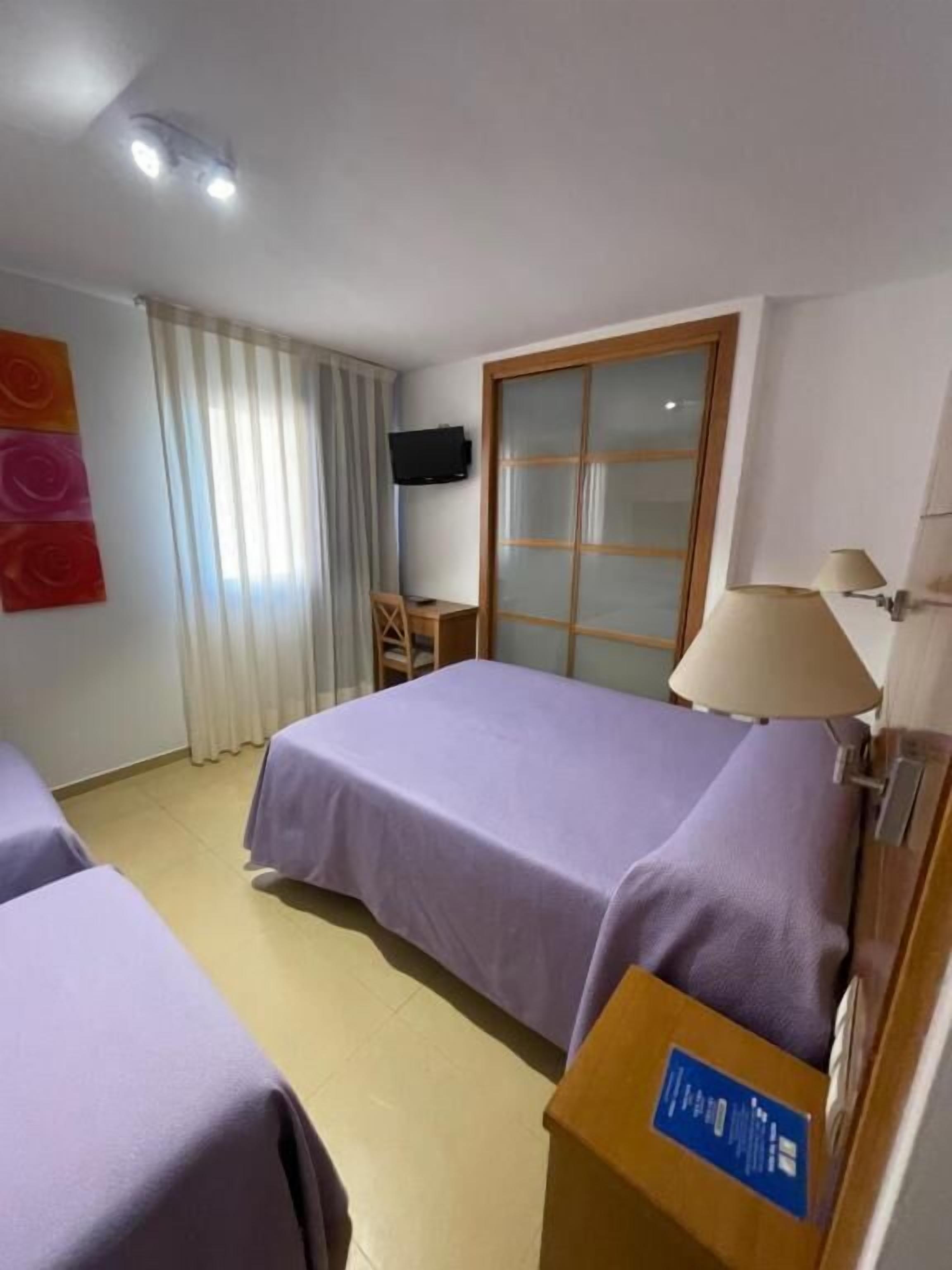 A hotel room with two beds, a TV, a desk, a chair, a window with curtains, and a sliding door.