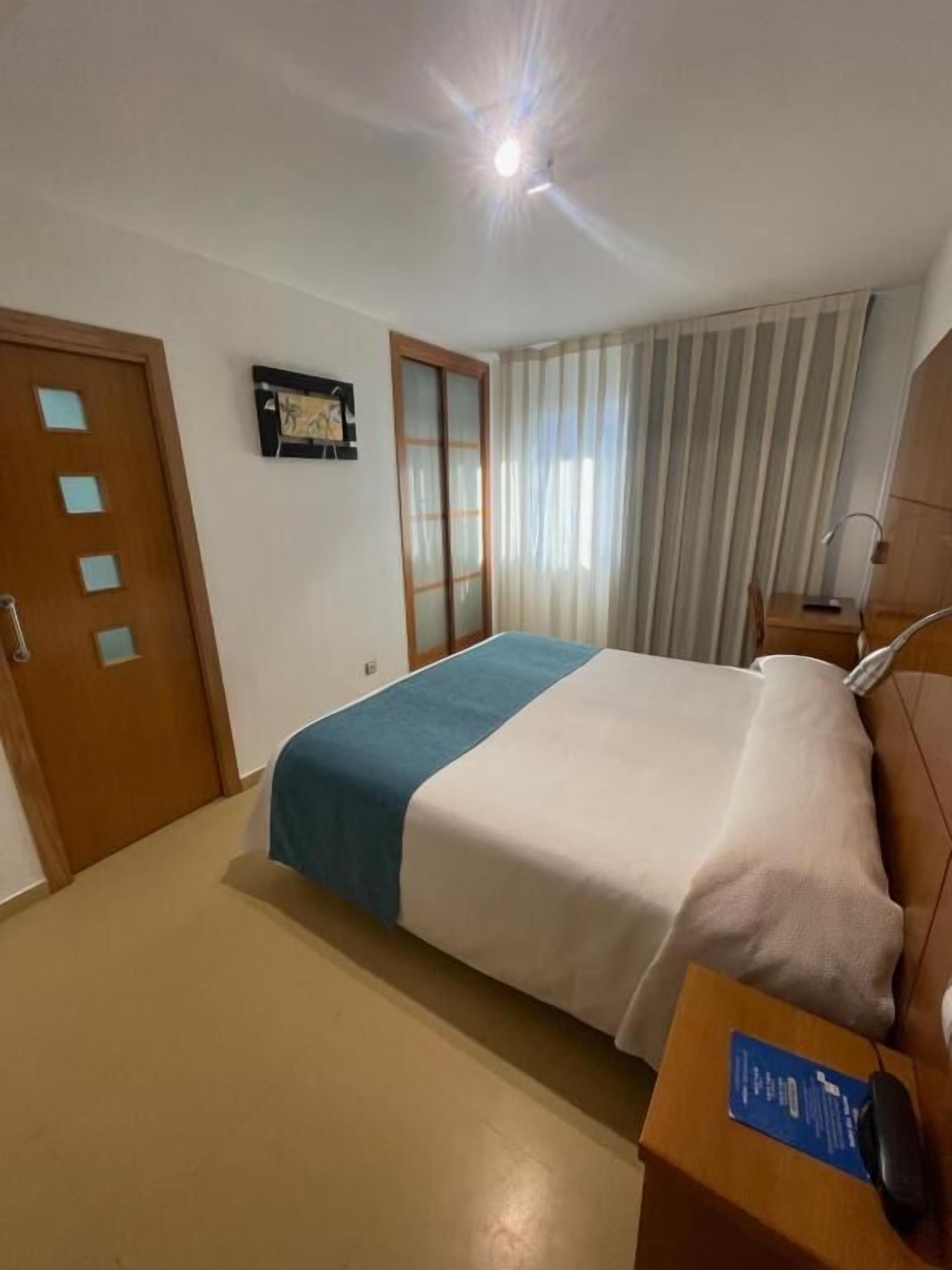 A hotel room with a bed, a desk with a computer, a chair, a door, and a window with curtains.