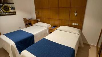 A hotel room with two single beds, a wooden headboard, a small table with a chair, and a painting on the wall.