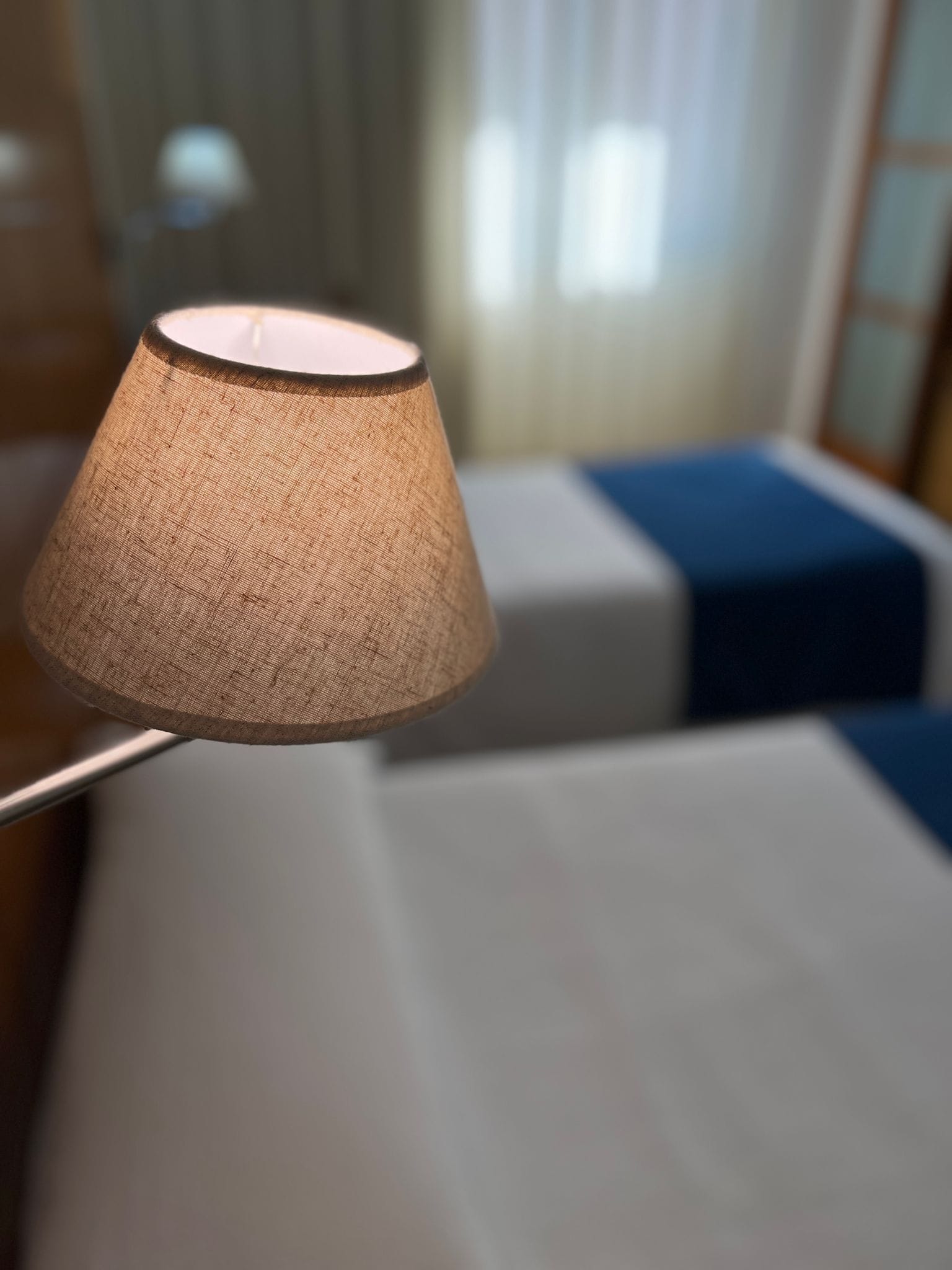 Primer plano de una lámpara con pantalla de tela texturizada, una cama con sábana blanca y una manta azul en una habitación de hotel.