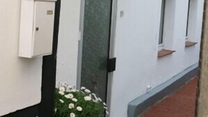 A narrow outdoor corridor with a white wall, a door with a frosted glass panel, a small potted plant with white flowers, and a red brick path. - Appartement Hexenhäuschen, 1-3 Pers (Haltern am See)