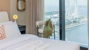 A hotel room with a large bed, a hanging lamp, a chair with a green throw, a mirror, and a view of a Ferris wheel through a large window. - Aya - Beachfront Bliss 2BR Haven at The Beach Residences (Dubai)