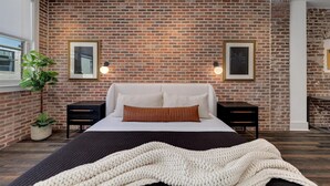 A bedroom with a brick wall, a bed with a white headboard, two bedside tables, and a large window. - Loft Style Spacious Lux Apt 202, sleeps 6 (Atlanta)