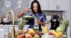 Une cuisine où une personne verse un liquide vert dans une bouteille, entourée de divers fruits et d’un appareil à jus.