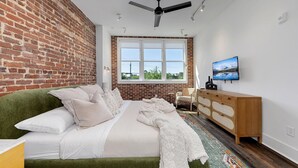 A bedroom with a brick wall, a green sofa, a wooden dresser, a television, and a large window with a view. - Loft Style Spacious Lux Apt 203, sleeps 6 (Atlanta)