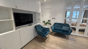 A modern living room with a flat-screen TV, two blue armchairs, a potted plant, and a sliding glass door leading to a bedroom. - Pop Art Luxury Room Marylebone  (London)