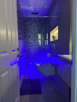 A narrow bathroom with a modern design, featuring a glass partition, a toilet, and a sink. - Pop Art Luxury Room Marylebone  (London)