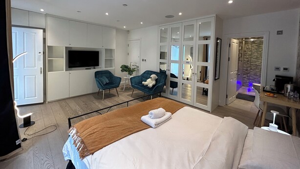 A modern hotel room with a bed, two armchairs, a TV, a wardrobe, and a bathroom with a shower. - Pop Art Luxury Room Marylebone  (London)