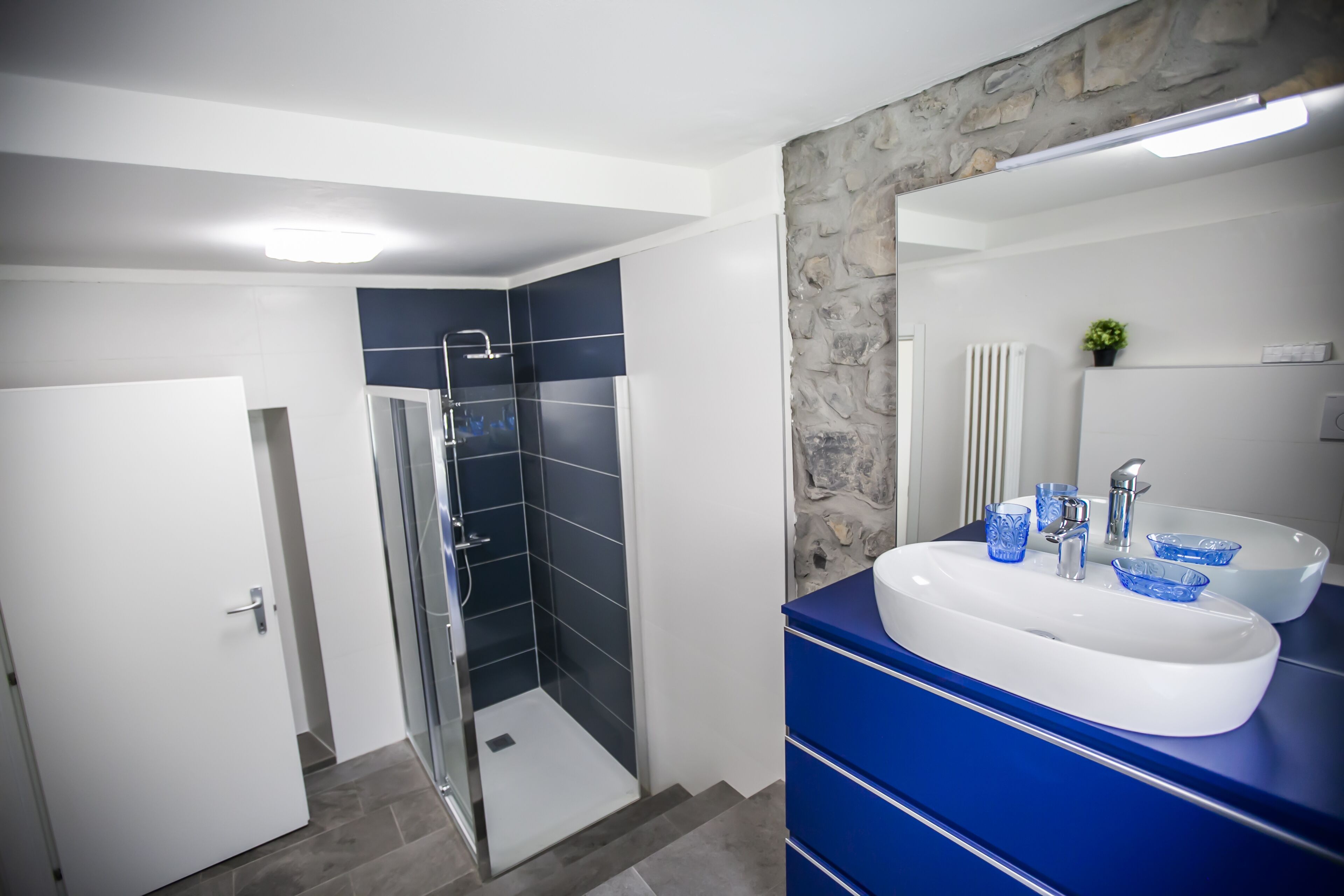 Un baño moderno con cabina de ducha de vidrio, un lavamanos blanco con cuencos de vidrio azules y un mueble de tocador azul.