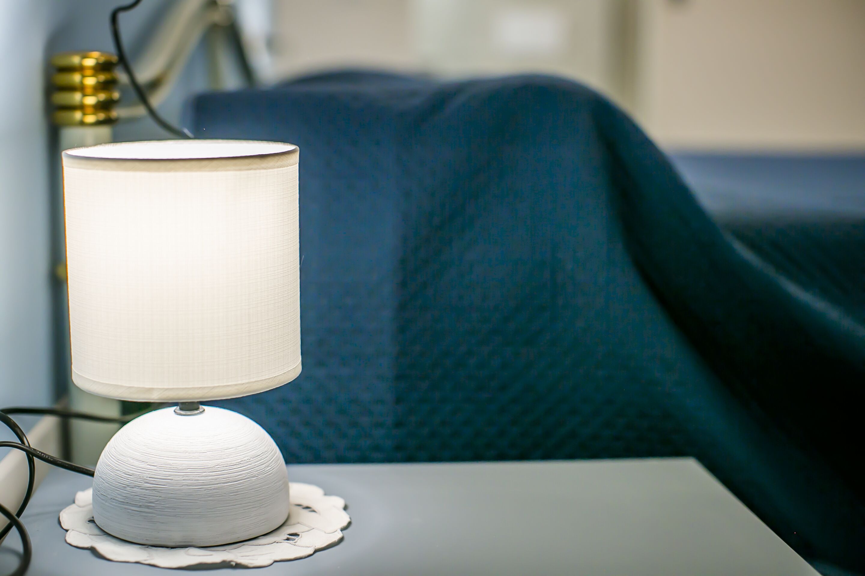 A white table lamp on a bedside table with a doily, a blue blanket, and a gold-colored lamp base.