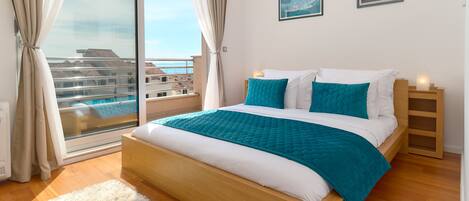 A bedroom with a large bed, a view of buildings through a sliding door, and a small bedside table with a lamp.