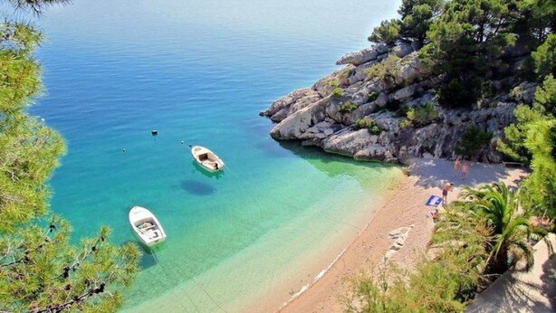 Une plage bordée d’une eau turquoise et limpide, d’une côte rocheuse et de quelques bateaux ancrés près du rivage.