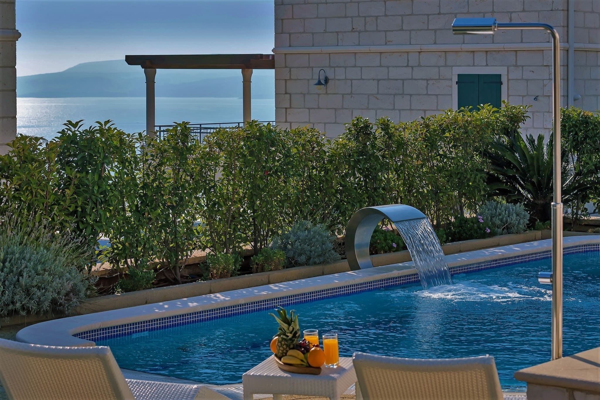 A pool with a waterfall feature, surrounded by lush greenery and a view of the sea.