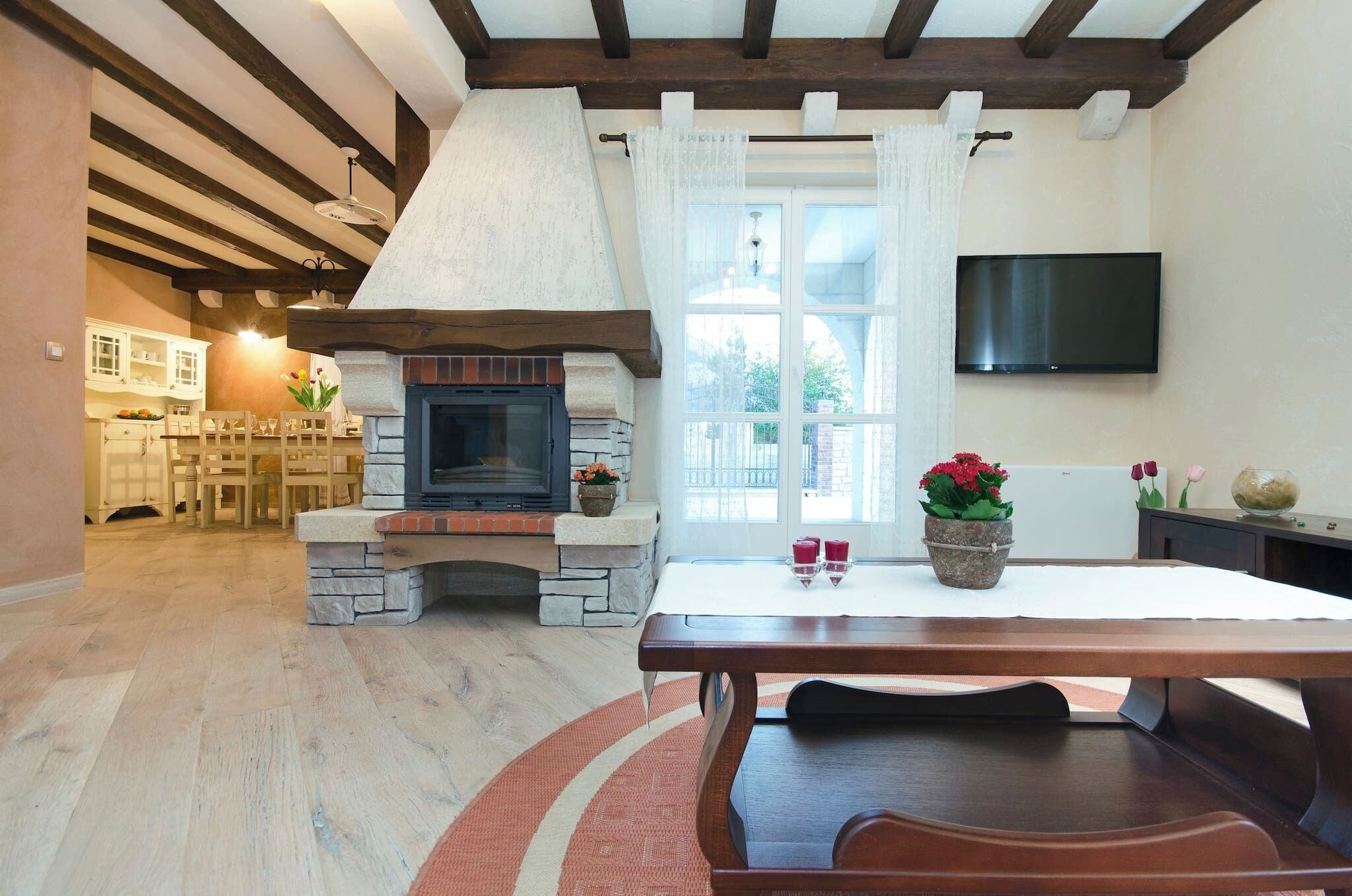 A cozy living room with a stone fireplace, wooden beams, a dining table, and a TV mounted on the wall.