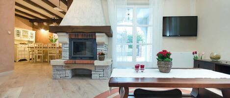 A cozy living room with a stone fireplace, wooden beams, a dining table, and a TV mounted on the wall.