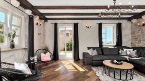 Un salon moderne avec un canapé foncé, une table basse ronde et un fauteuil noir. Une grande fenêtre avec des rideaux, un lustre et une vue sur l’extérieur complètent l’ensemble.