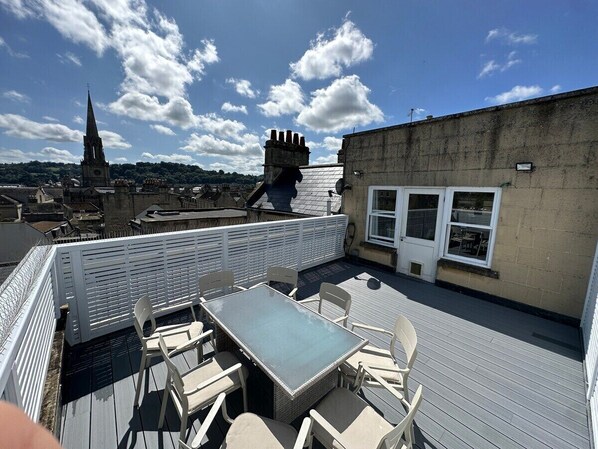 A rooftop terrace with a table and chairs, a white fence, and a view of a church spire in the background.