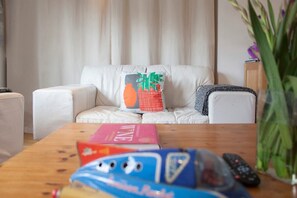 Un salon avec un canapé blanc, une table basse sur laquelle sont posés des livres et une télécommande, et un vase contenant des plantes vertes.