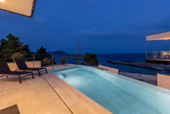 A modern outdoor swimming pool with lounge chairs, a view of the sea, and a mountainous landscape in the background. - Luxury Villa Luce, right on the beach (Potomje)