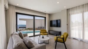 Ein modernes Wohnzimmer mit einer Couch, zwei gelben Sesseln, einem Couchtisch und einem Fernseher. Große Fenster bieten Blick auf einen Pool und das Meer.
