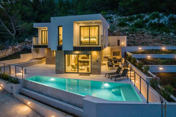 A modern two-story house with a swimming pool, outdoor seating, and a well-lit interior. - Luxury Villa Luce, right on the beach (Potomje)