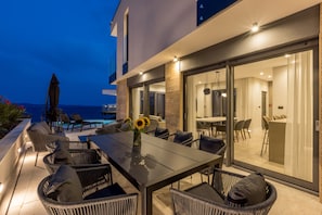 An outdoor dining area with wicker furniture and a table, overlooking a pool and a building with large glass doors. - Luxury Villa Luce, right on the beach (Potomje)