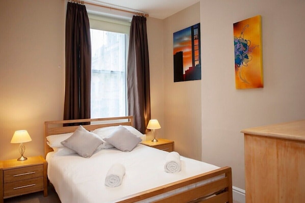 Quarto de hotel com uma cama, duas luminárias de cabeceira, uma mesa de cabeceira de madeira, um penteou de madeira, uma janela com cortinas e duas obras de arte na parede.