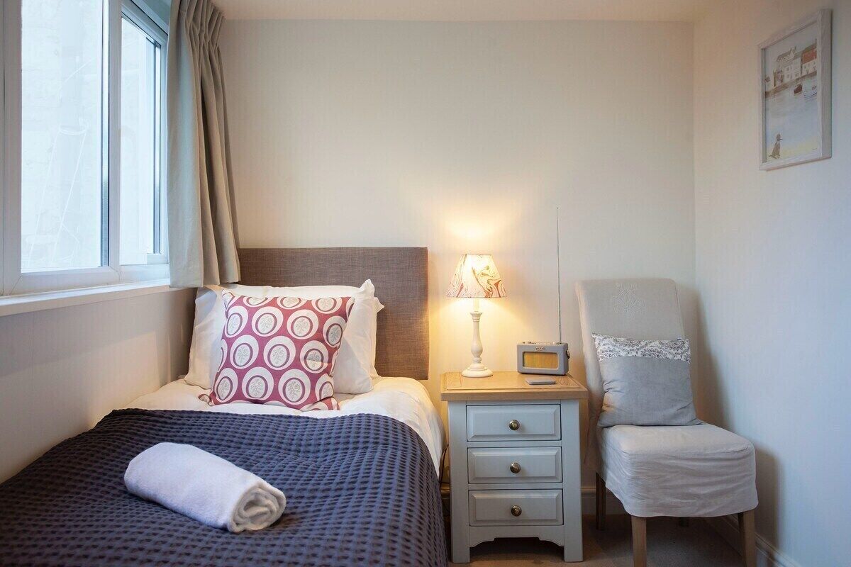 A neatly made bed with a patterned pillow, a bedside lamp, a chair with a lace cover, and a small table with a radio.