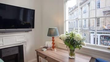 A room with a television mounted on the wall, a fireplace, a table with a lamp and a vase of flowers, and a window with a view of buildings.
