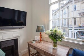 A room with a television mounted on the wall, a fireplace, a table with a lamp and a vase of flowers, and a window with a view of buildings.