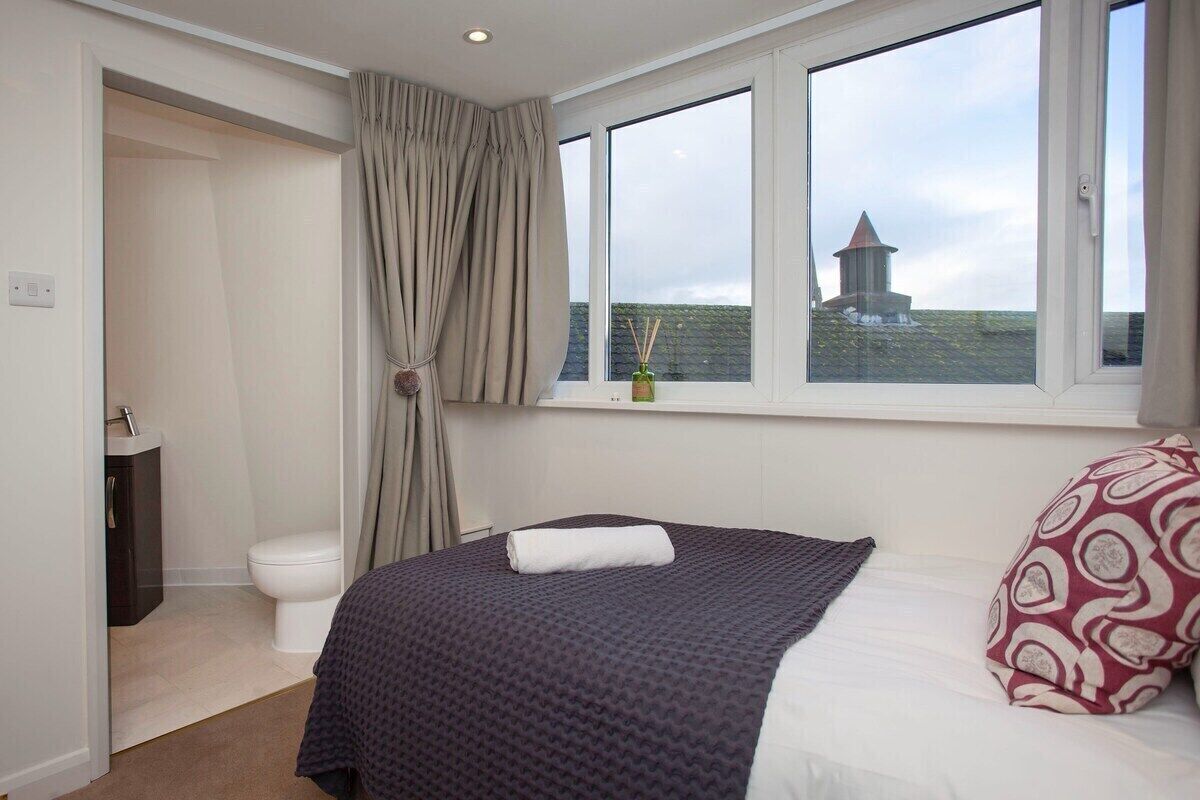 A bedroom with a large window, a bed with a patterned pillow, a bedside table, and a view of a lighthouse.