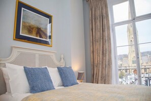 Une chambre d’hôtel avec un lit, un tableau encadré, une fenêtre avec des rideaux et une vue sur un bâtiment à l’extérieur.